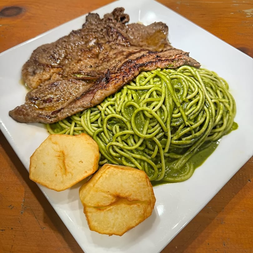 Peruvian pesto spaghetti with grilled steak and two fried potatoes on a white plate.
